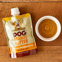 Thumbnail for A pouch of Peanut Butter Dog food next to a small white bowl filled with peanut butter, set on a wooden tabletop.