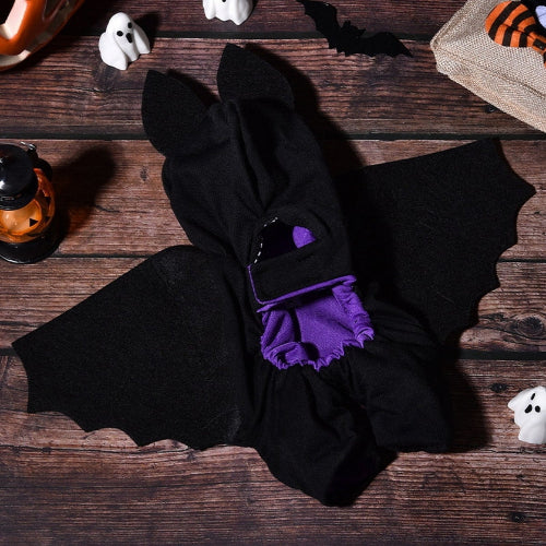 A black bat costume laid flat on a dark wooden surface, featuring pointed ears, bat wings, and a purple ruffled collar detail. Halloween props including small ghost decorations, a jack-o'-lantern lantern, and a burlap sack are arranged around it.