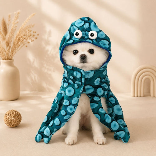 A small white dog wearing a blue octopus-themed hooded pet blanket with large cartoon eyes on the hood, sitting on a beige surface in a styled indoor setting.