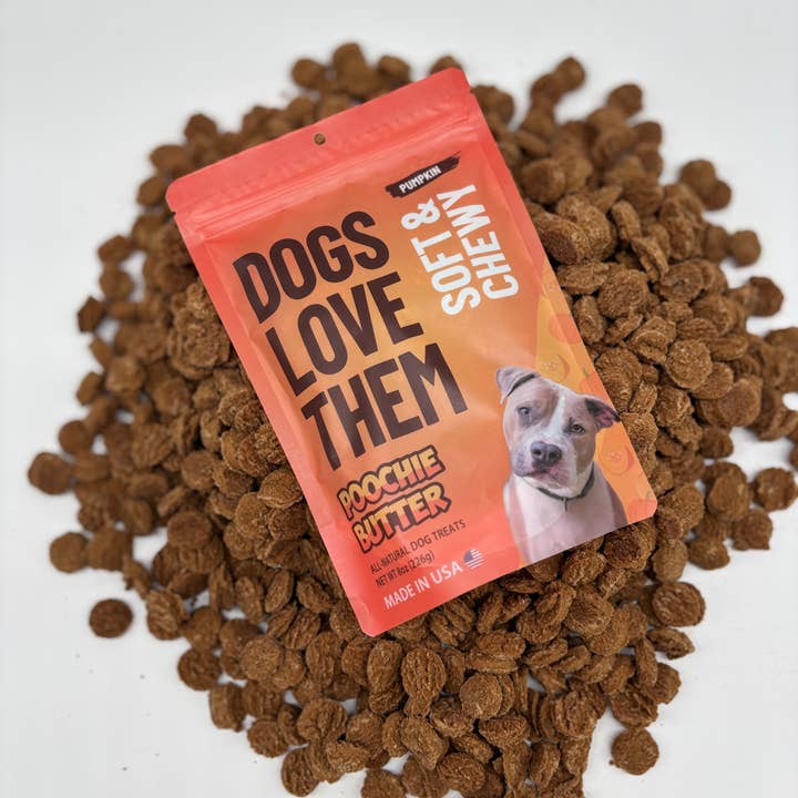 A bag of Poochie Butter dog treats placed on top of a pile of brown kibble treats on a white background. The bag is orange and pink with black text reading 'DOGS LOVE THEM' and 'SOFT & CHEW'.