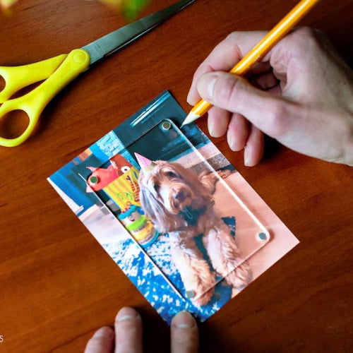 A clear acrylic photo frame being held on a wooden table by a person's hands, with a printed pet photo inside the frame and yellow-handled scissors nearby.