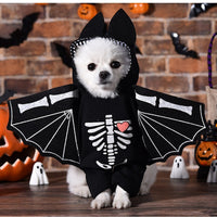 Thumbnail for A small white dog wearing a black bat costume with silver glitter wings and a white skeleton print with a pink heart, posed in a Halloween-themed setting with pumpkins, ghosts, and a brick wall background.